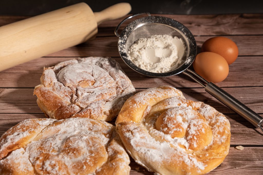 Ensaimada de mallorca. Tres ensaimadas de Mallorca (enseimadas), sobre una tabla de madera, con azúcar para espolvorear, huevos y un rodillo para amasar. Dulce típico de la gastronomía mallorquina.